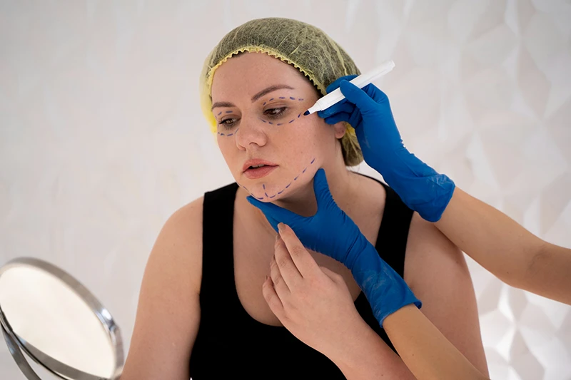 Young woman with cosmetic surgery markings examining her face in a mirror during consultation with a doctor