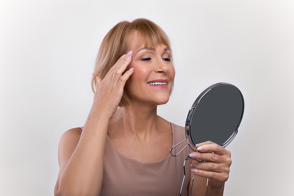 Smiling woman looking in a mirror and touching her face, showing satisfaction with mini facelift results